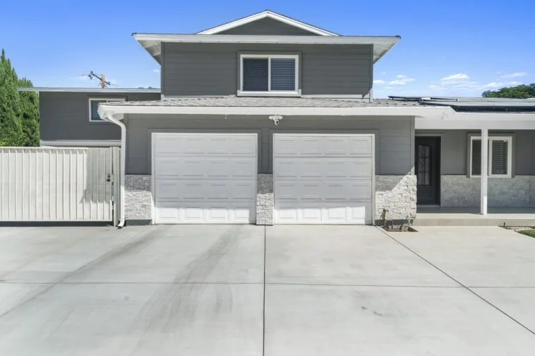 Two-story gray home exterior with dual white garage doors and stacked stone veneer wainscoting by RnD Builders Inc