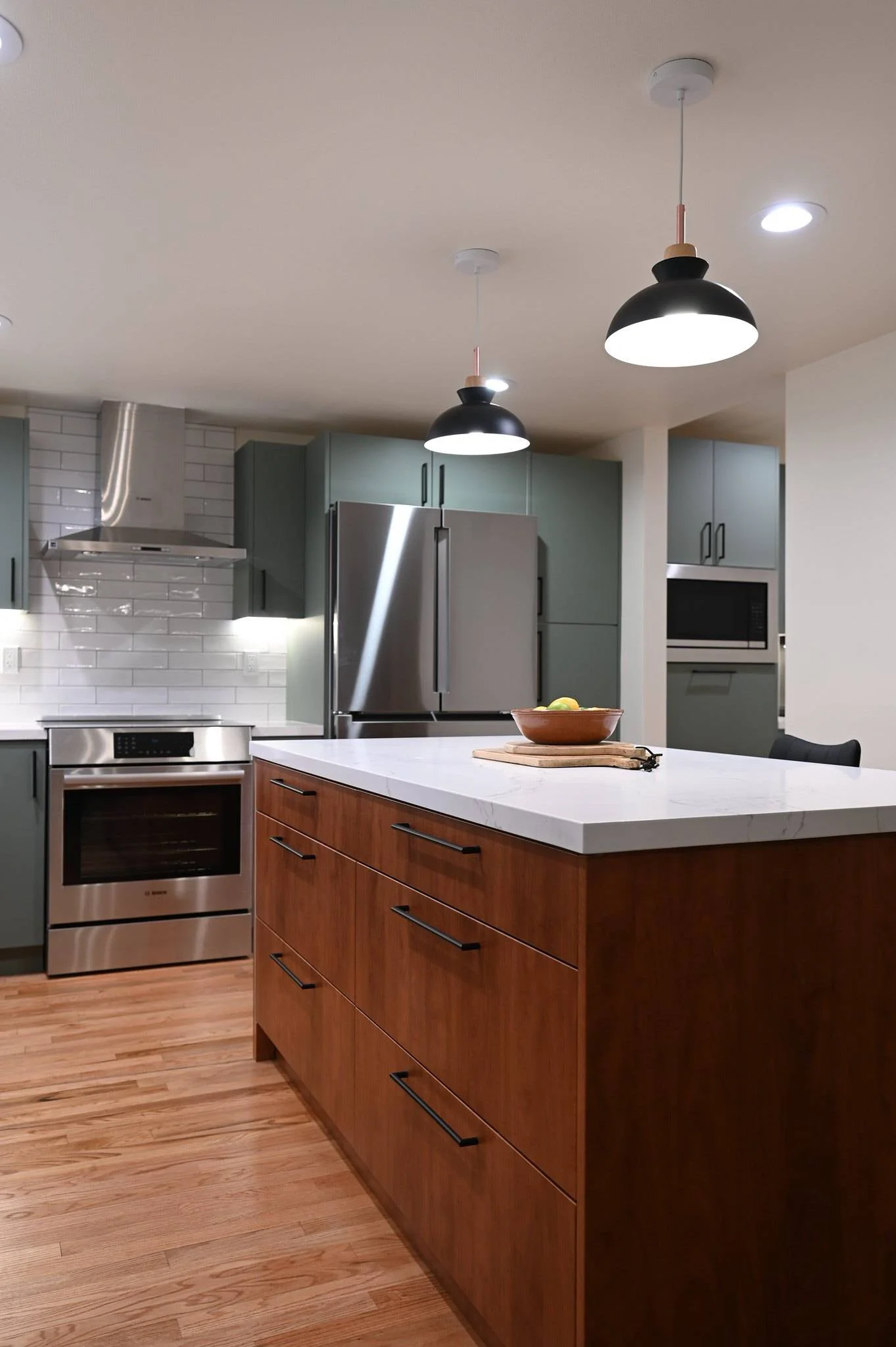 Modern kitchen with walnut island, quartz countertop, sage green cabinets, and black pendant lights by RnD Builders Inc