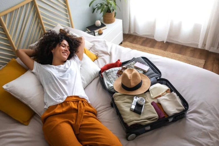 Woman relaxing on a bed beside an open packed suitcase in a bright bedroom by RnD Builders Inc