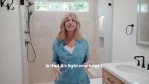 Woman presenting a bright remodeled bathroom with walk-in shower and matte black fixtures by RnD Builders Inc