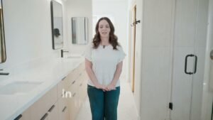 Woman standing in modern bathroom with light wood vanity, white quartz countertop, and frameless glass shower by RnD Builders Inc