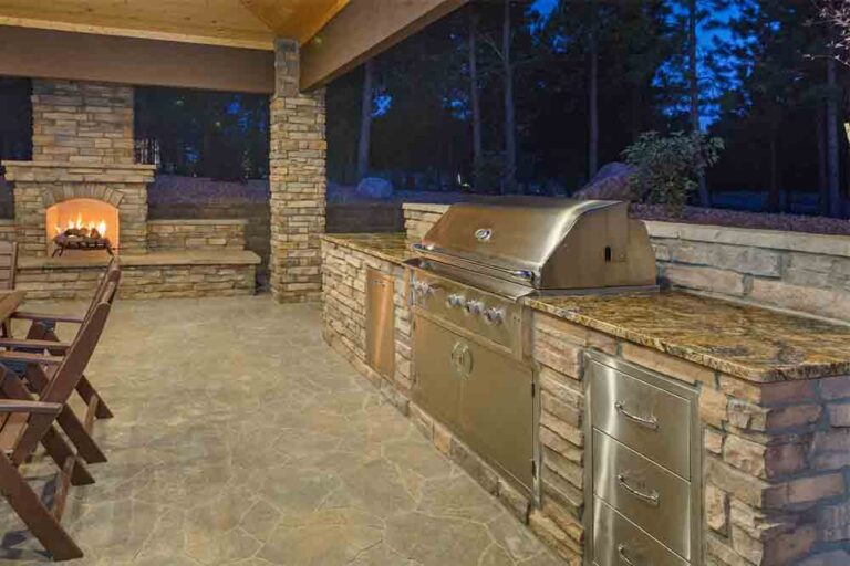 Covered outdoor kitchen with stainless steel grill, granite countertop, stacked stone surround, and lit fireplace by RnD Builders Inc