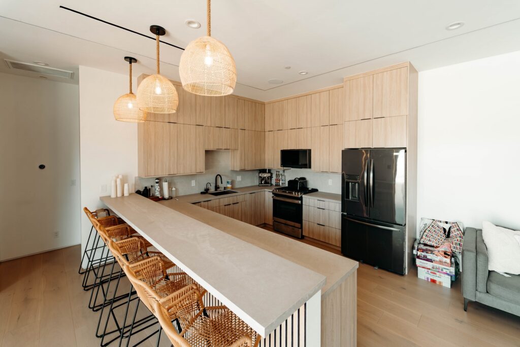 Modern kitchen with light oak flat-panel cabinets, stone peninsula, and woven rattan pendant lights by RnD Builders Inc