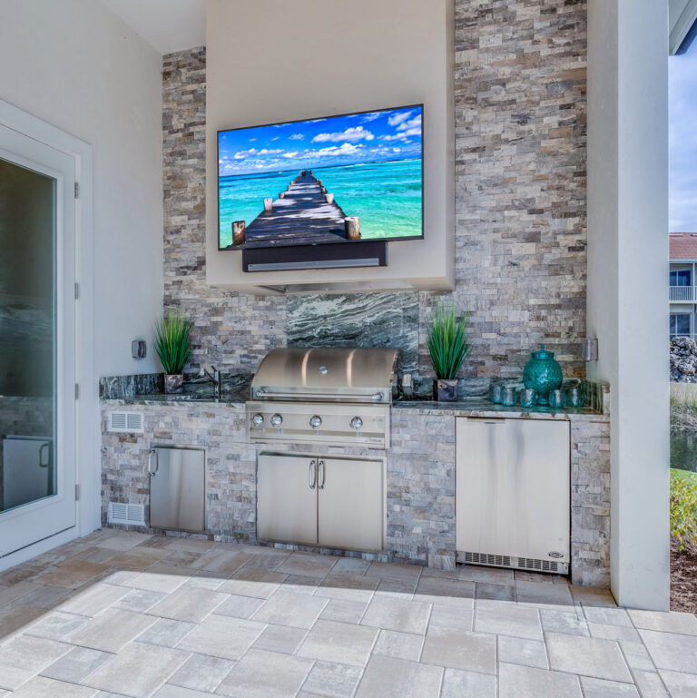 Covered outdoor kitchen with stainless steel grill, undercounter fridge, stone veneer, and wall-mounted TV by RnD Builders Inc