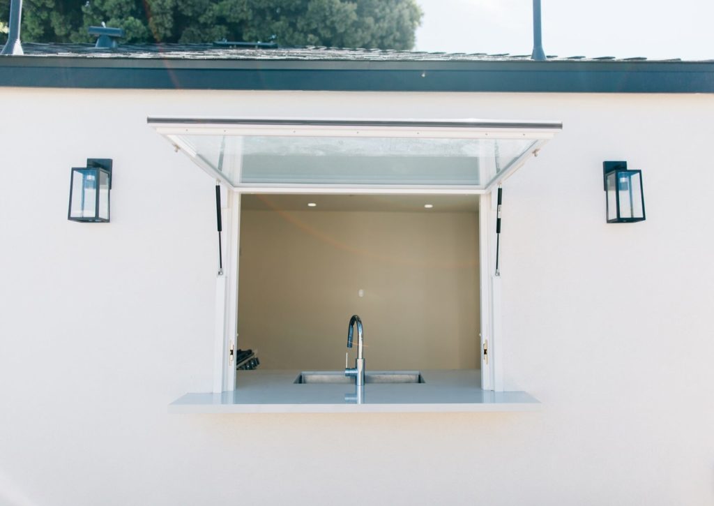 Exterior Pass-through Window with Glass Awning Exterior pass-through window with glass awning, undermount sink, and black wall sconces by RnD Builders Inc