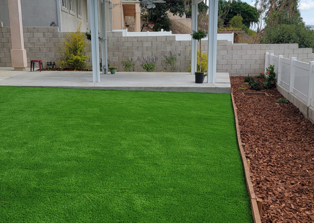 Backyard Landscaping with Artificial Turf Backyard landscaping with artificial turf, concrete patio, wood-framed mulch planting bed, and white pergola by RnD Builders Inc