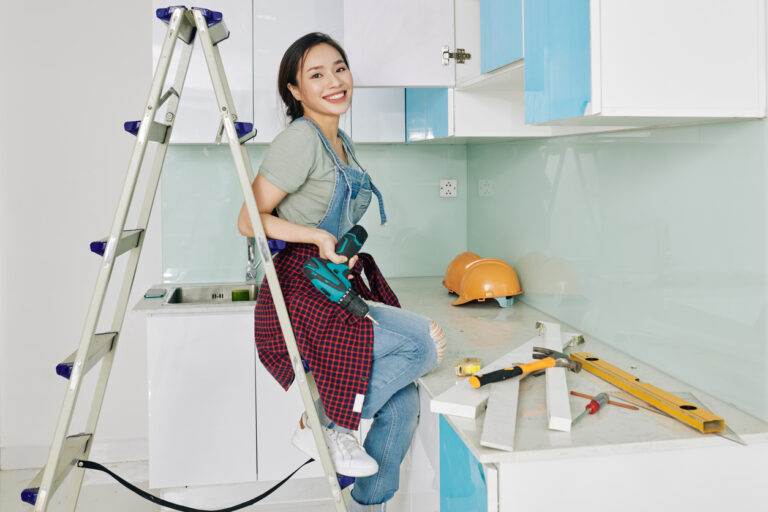 A woman sits on a ladder in a kitchen, surrounded by cabinets and cooking utensils, focused on a task above her.