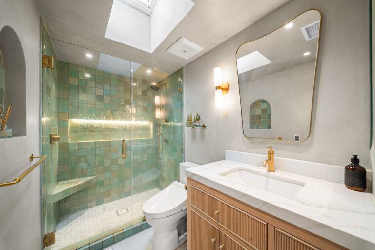Green Tile Shower and Wood Vanity with a green tile shower, a light wood vanity, a shaped mirror and brass fixtures in Manhattan Beach by RnD Builders Inc