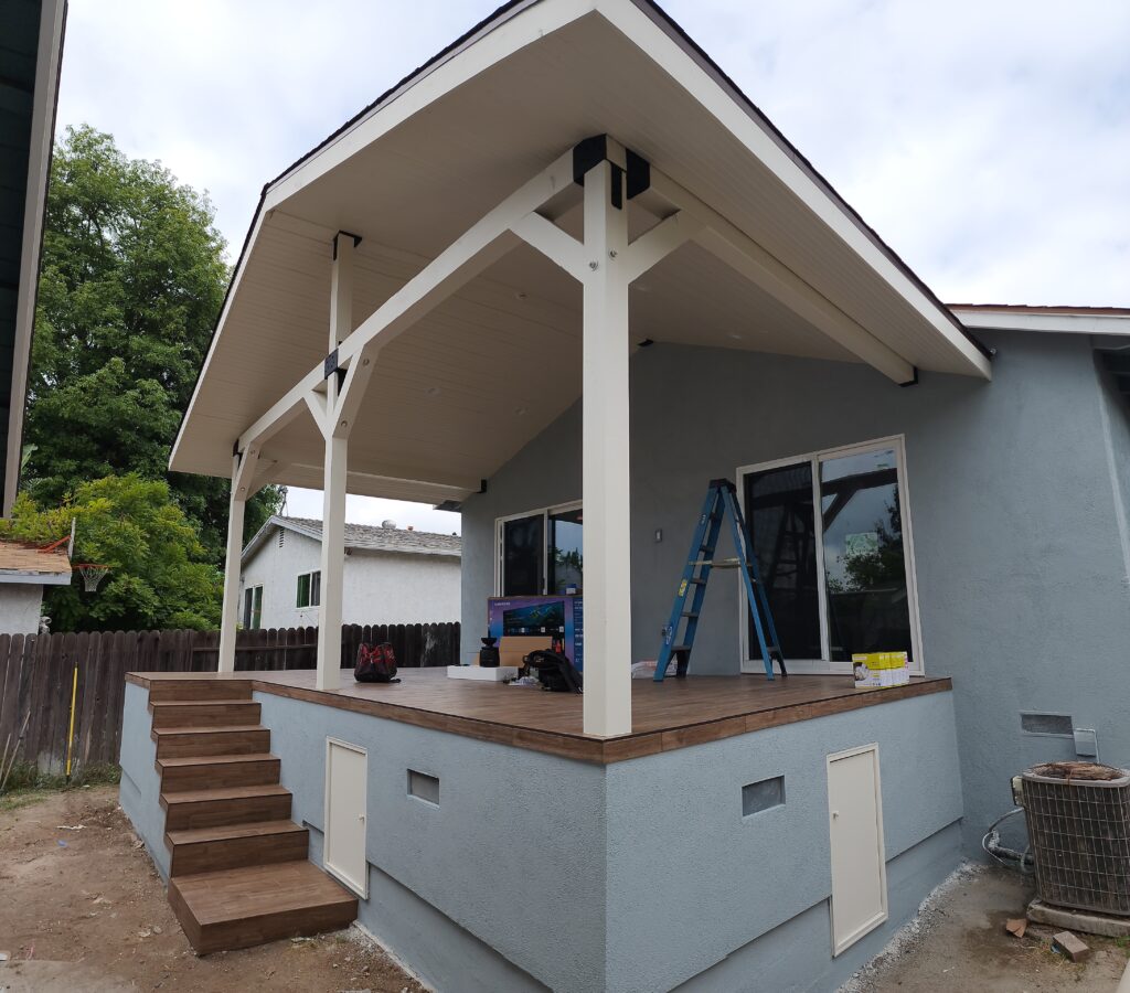 Covered gable-roof patio addition with wood deck and white painted timber posts by RnD Builders Inc