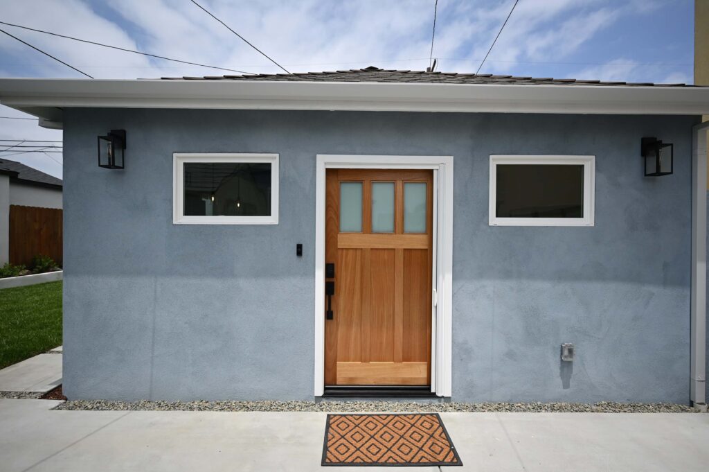 Hawthorne 750 sqft ADU: Hawthorne 750 sqft ADU: ADU exterior with blue-gray stucco, craftsman wood door, and black wall sconces in Hawthorne by RnD Builders Inc