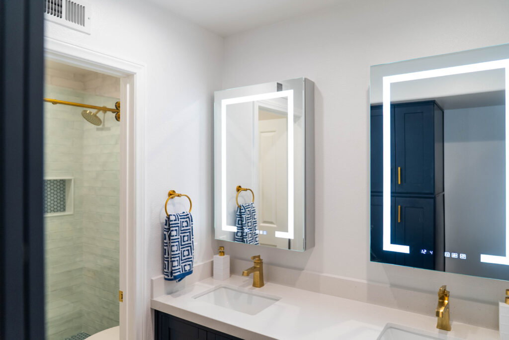 Oxnard bathroom remodel: Modern bathroom with navy vanity, LED mirror cabinet, gold fixtures, and tiled shower in Oxnard by RnD Builders Inc