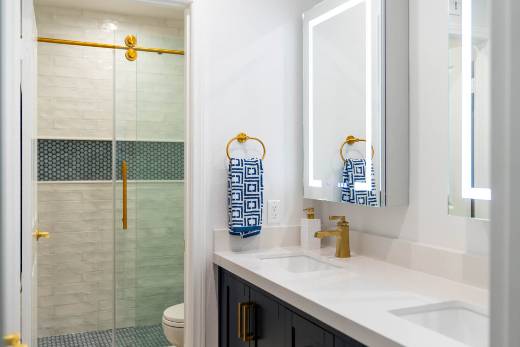 Modern bathroom with dark navy vanity, white quartz countertop, LED mirror, and glass shower with brushed gold fixtures by RnD Builders Inc