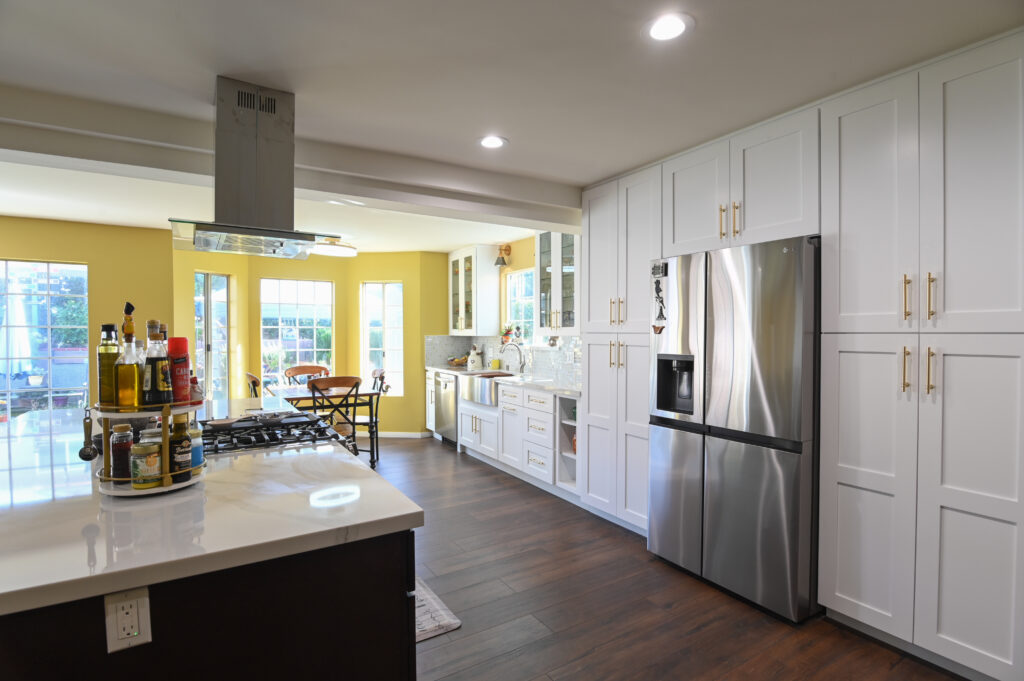 Open-concept kitchen with white shaker cabinets, stainless steel refrigerator, and dark hardwood floors by RnD Builders Inc