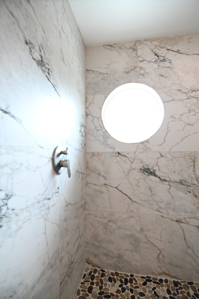 Moorpark bathroom remodel: Walk-in shower with large-format marble tile walls, pebble stone floor, and round porthole window in Moorpark by RnD Builders Inc