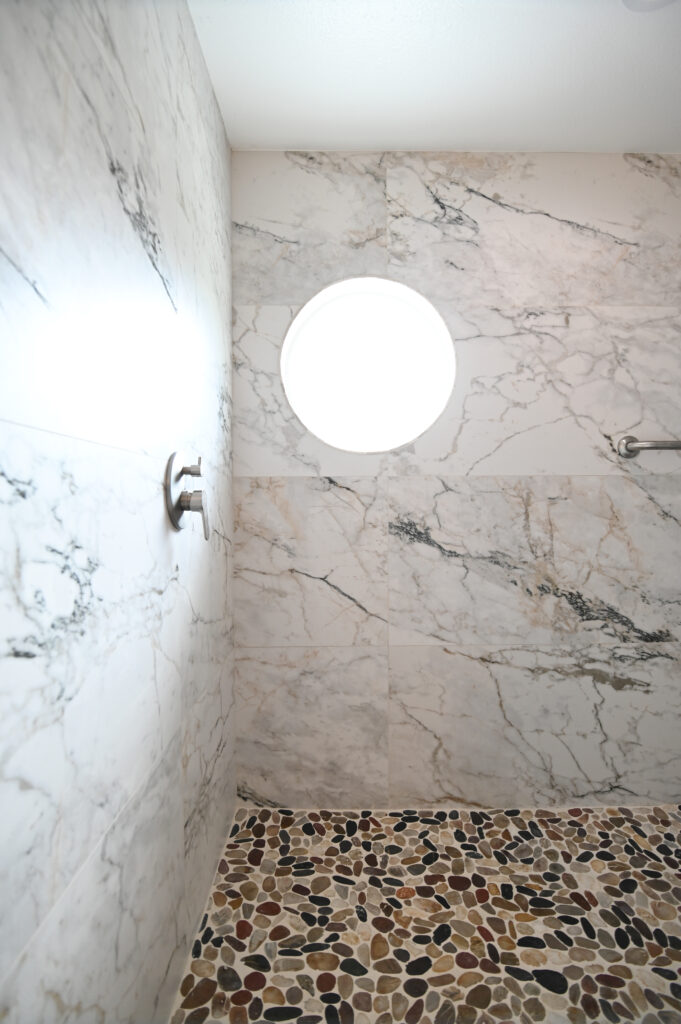 Moorpark bathroom remodel: Walk-in shower with large-format marble wall tiles, pebble stone floor, and circular porthole window in Moorpark by RnD Builders Inc