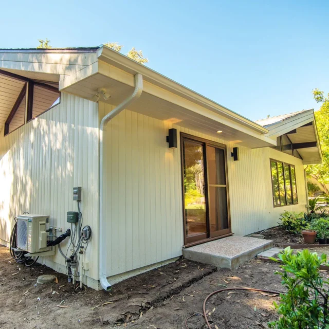Exterior Addition with Cream Vertical Board Siding Exterior addition with cream vertical board siding, sliding glass door, and mini-split HVAC unit by RnD Builders Inc