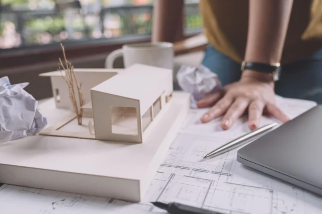 Architectural model of a house, blueprints, and a laptop, on a desk. Architectural plans by RnD Builders