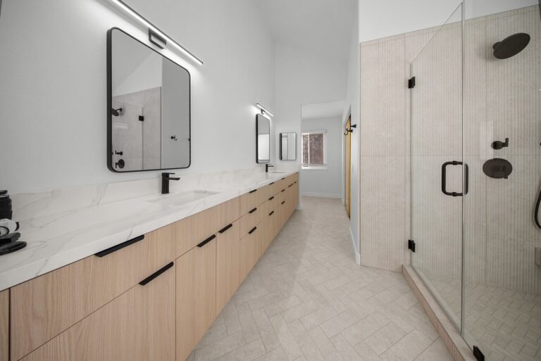 Modern Primary Bathroom with Long Light Wood Vanity Modern primary bathroom with long light wood vanity, white quartz countertop, and glass shower enclosure by RnD Builders Inc